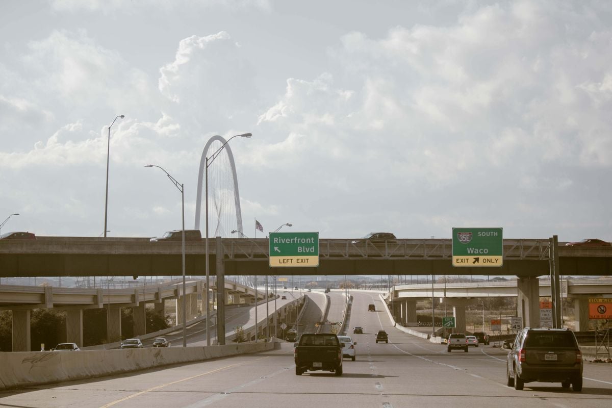 A Dallas expressway with the Margaret Hunt Hill Bridge arch and highway signs for I-35E, with cars heading out of the city