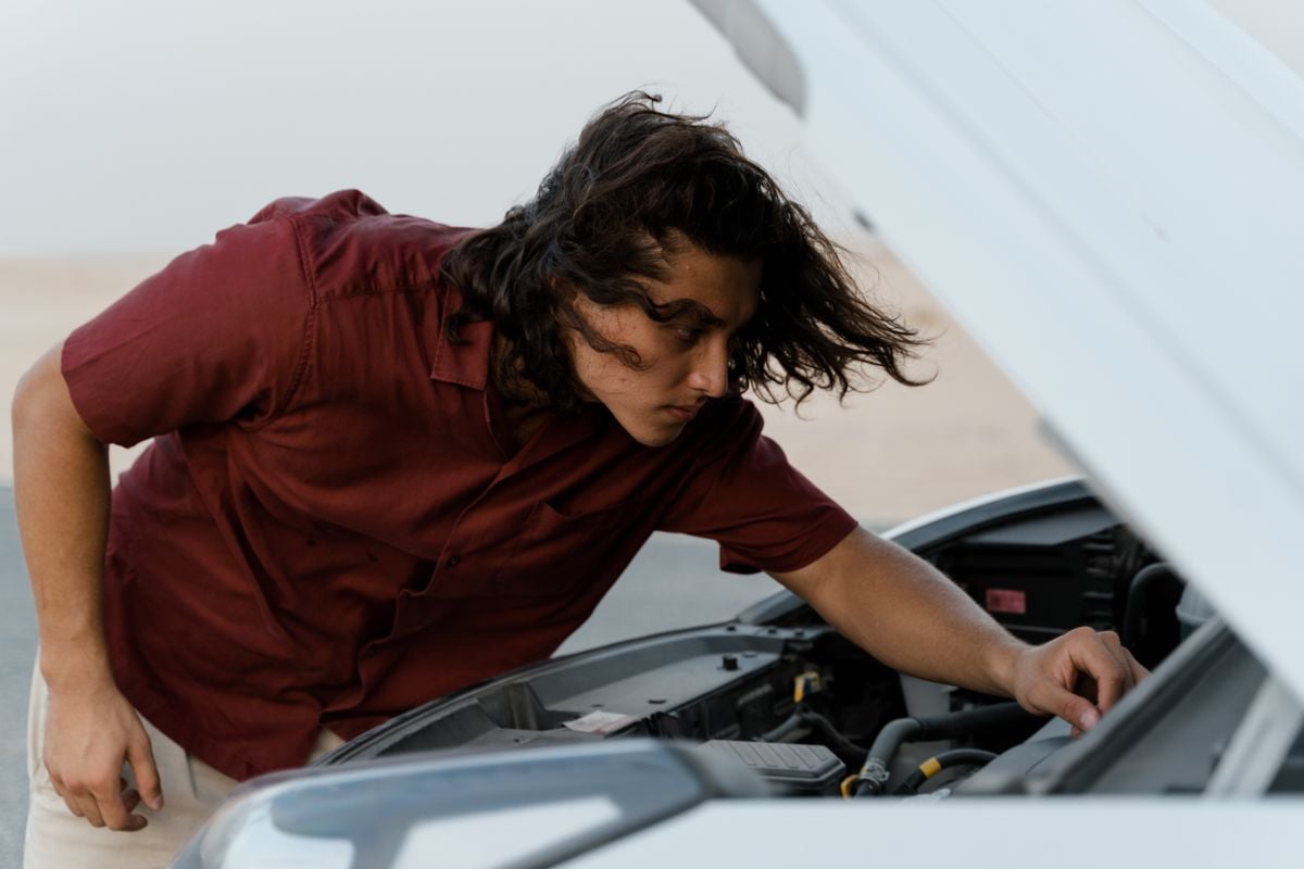 A young man inspecting the engine bay of a used car under the raised hood on a bright day